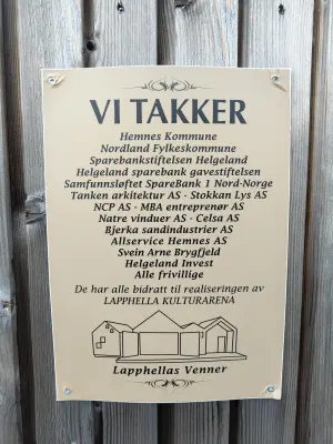 Et skilt med teksten 'VI TAKKER' som uttrykker takknemlighet til ulike bidragsytere for realiseringen av Lapphella Kulturarena på Hemnesberget. Skiltet lister opp kommunale, regionale, private og frivillige aktører, og er montert på en trevegg. Nederst vises en enkel strekillustrasjon av kulturarenaen og teksten 'Lapphellas Venner'.
